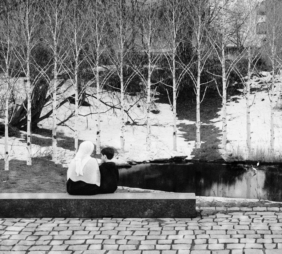 Sort-hvitt bilde av trær, et vann og to mennesker som sitter på en benk laget av stein
